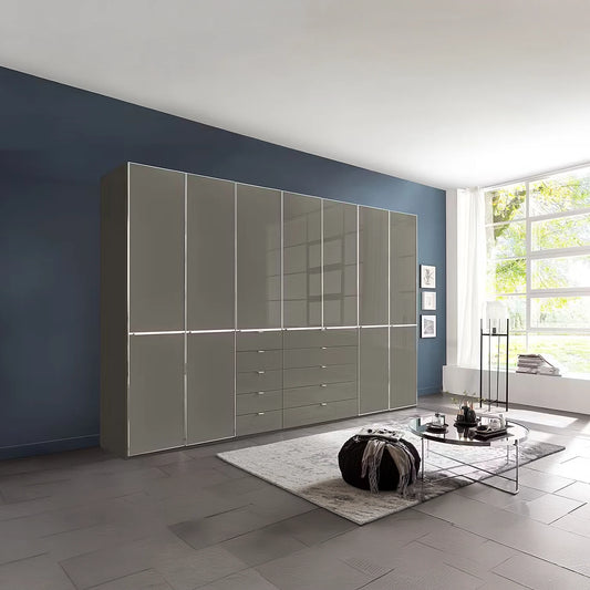 A large modern Wiemann Santa 350cm Combi 7 Doors Wardrobe With Drawers in Havana. The cabinet features glossy Havana Glass doors and drawers with slim silver handles, set against a dark blue accent wall.