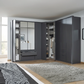 A modern dark gray corner wardrobe system in a bedroom, shown with doors open to reveal organized shelves, hangers, and folded clothes. The room includes a bed with a black-and-white patterned duvet, a gray shag rug, light wood flooring, and large windows on the left.