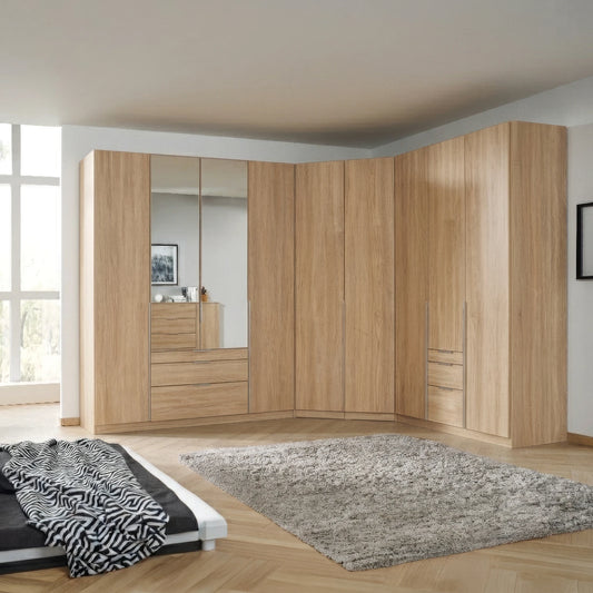 Rauch Alabama large curved corner wardrobe system in a light oak wood finish, featuring multiple doors, drawers, and mirrored panels, shown in a modern bedroom setting with a gray shag rug and a bed partially visible in the foreground.