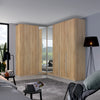 A large corner wardrobe set in a modern bedroom, featuring light oak wood grain doors with mirrored panels on the inner doors to show the bed underneath.