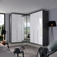 A modern Rauch Alabama corner wardrobe with white high-gloss doors and dark gray side panels, including mirrored doors that reflect the room. The wardrobe is installed in a contemporary bedroom setting with a bed, side tables, and a floor lamp.