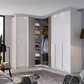 A modern corner wardrobe system in a bright, contemporary bedroom. The system features two tall white doors on the right, two white doors on the left, and an open center section showing shelves, hangers, and folded clothes.