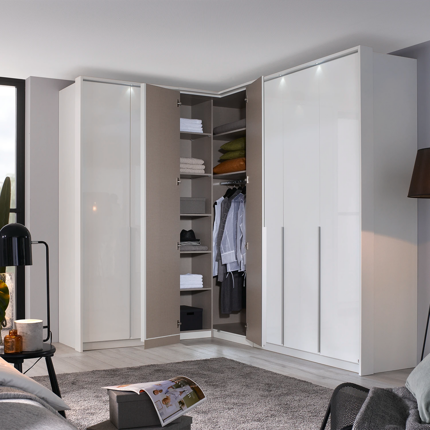 A modern corner wardrobe system in a bright, contemporary bedroom. The system features two tall white doors on the right, two white doors on the left, and an open center section showing shelves, hangers, and folded clothes.