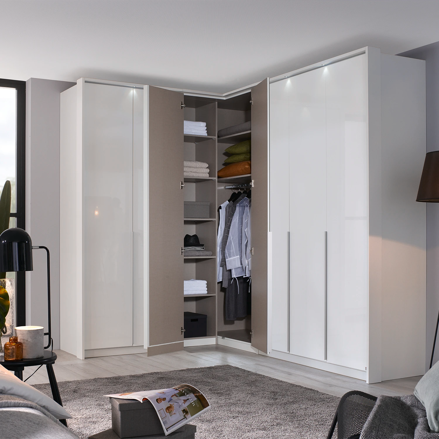 A modern corner wardrobe system in a bright, contemporary bedroom. The system features two tall white doors on the right, two white doors on the left, and an open center section showing shelves, hangers, and folded clothes.