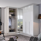 A modern white wardrobe with sliding glass doors and open interior shelving and hanging space, shown in a styled bedroom setting with a rug, lamp, and window view.