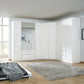 A large white corner Rauch Alabama  wardrobe system in a modern bedroom, featuring solid doors, mirrored panels, and built-in drawers, set against light wood flooring and a gray shag rug.
