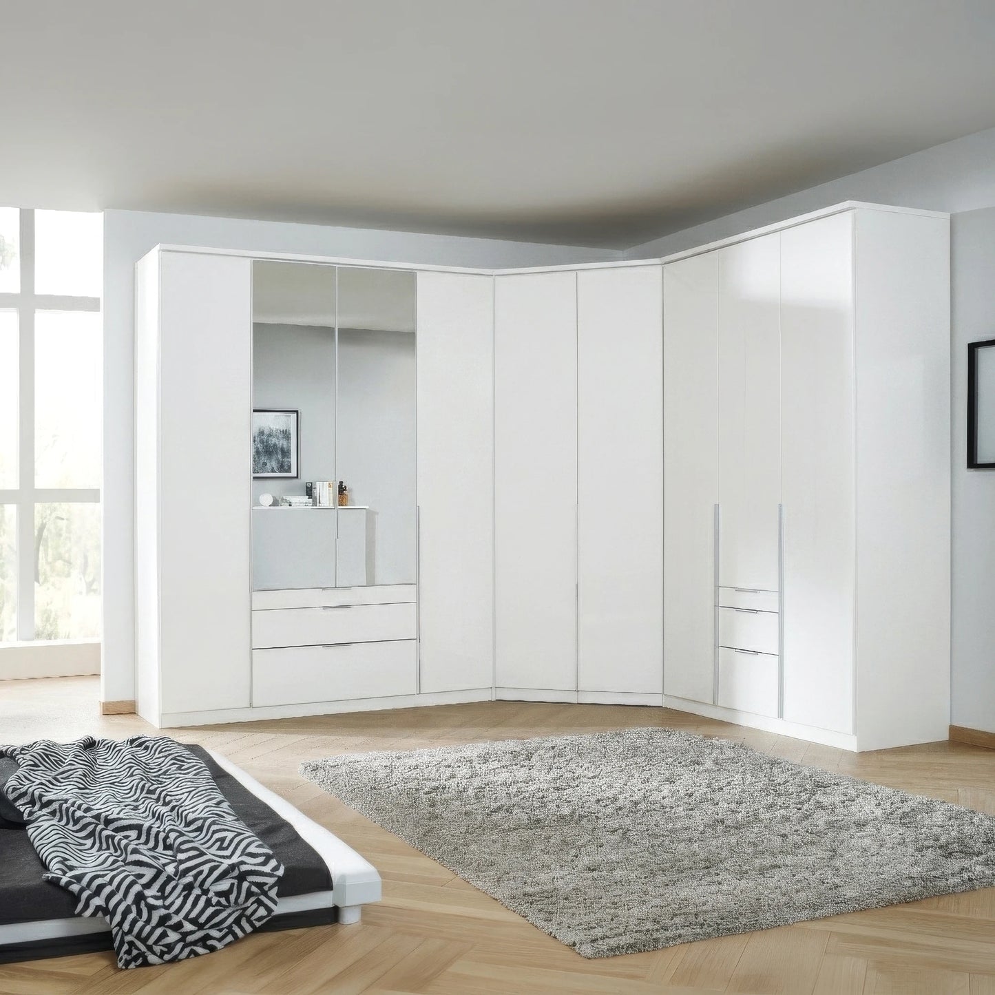 A large white corner Rauch Alabama  wardrobe system in a modern bedroom, featuring solid doors, mirrored panels, and built-in drawers, set against light wood flooring and a gray shag rug.