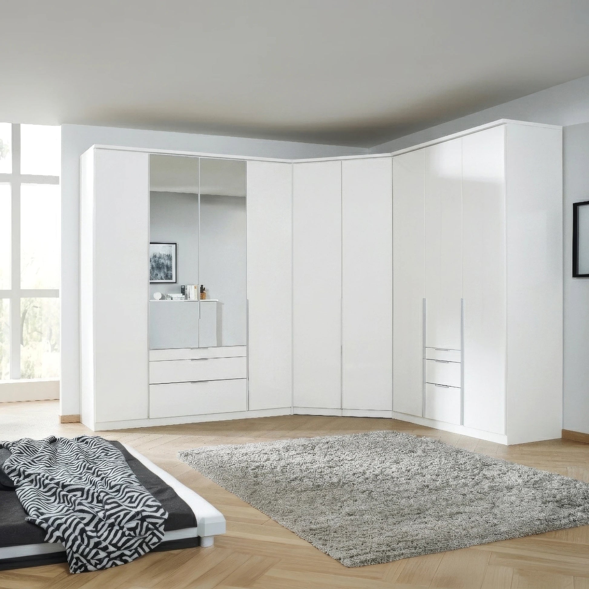 A large white corner Rauch Alabama  wardrobe system in a modern bedroom, featuring solid doors, mirrored panels, and built-in drawers, set against light wood flooring and a gray shag rug.