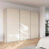 A modern Rauch Impulse three-door sliding wardrobe in a warm Champagne finish, set in a brightly lit bedroom with light wood floors and a white bed visible in the foreground.