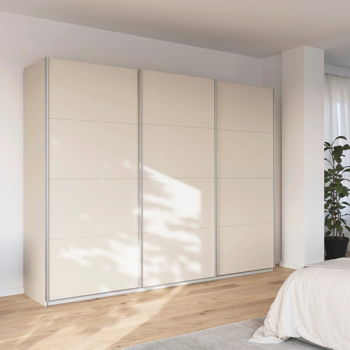 A modern Rauch Impulse three-door sliding wardrobe in a warm Champagne finish, set in a brightly lit bedroom with light wood floors and a white bed visible in the foreground.