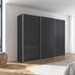 A modern three-door sliding rauch impulse wardrobe in a dark charcoal gray finish with slim silver metal frames, set against a white wall in a bright, contemporary bedroom.