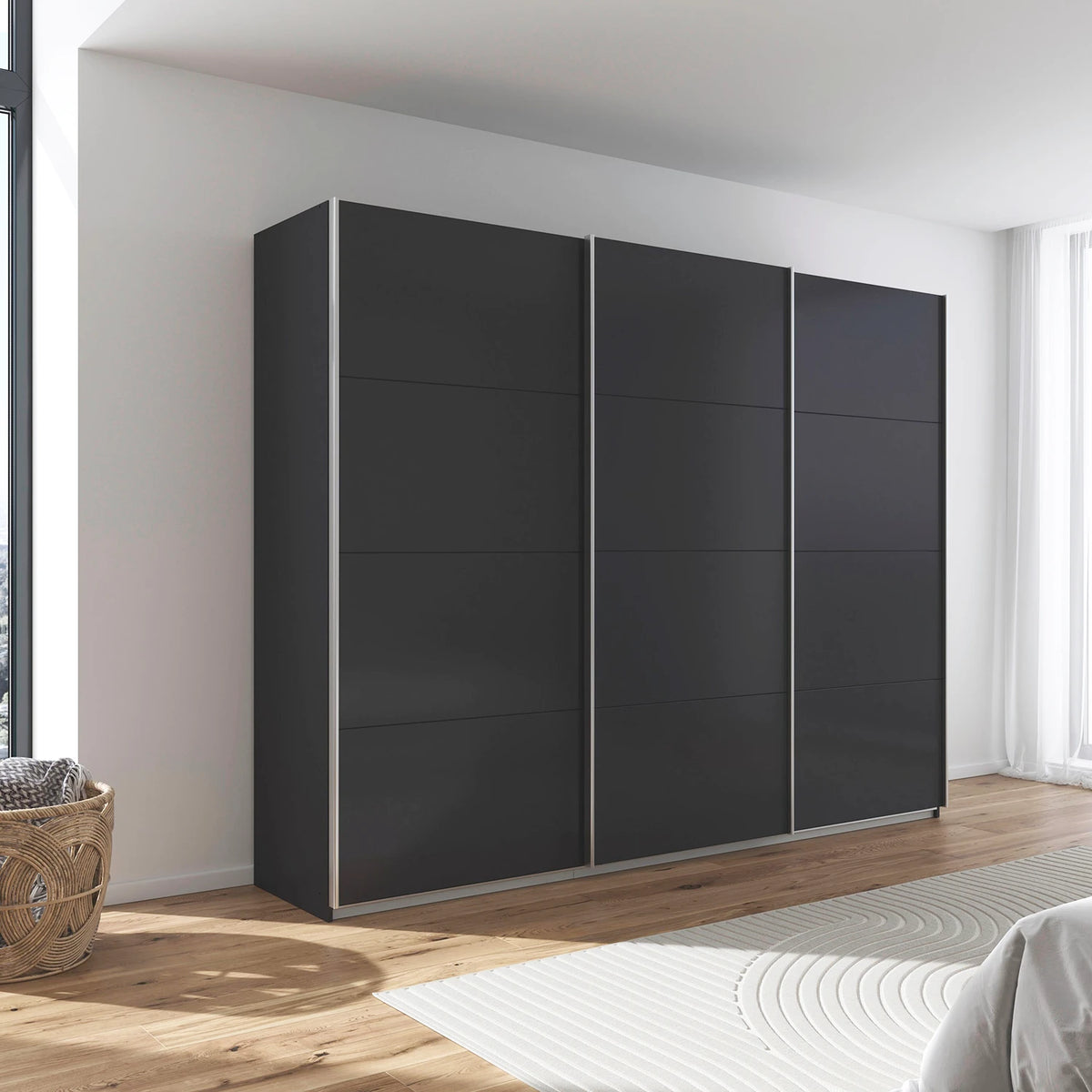 A modern three-door sliding rauch impulse wardrobe in a dark charcoal gray finish with slim silver metal frames, set against a white wall in a bright, contemporary bedroom.