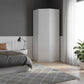 A tall white corner wardrobe in a modern bedroom setting with a gray wall, white wainscoting, a bed, a side table with a black lamp, sheer white curtains, and a potted green plant.