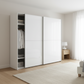 A modern white sliding wardrobe with one open door showing hanging clothes and shelves, set in a bright, minimalist bedroom with light wood flooring and a beige rug.