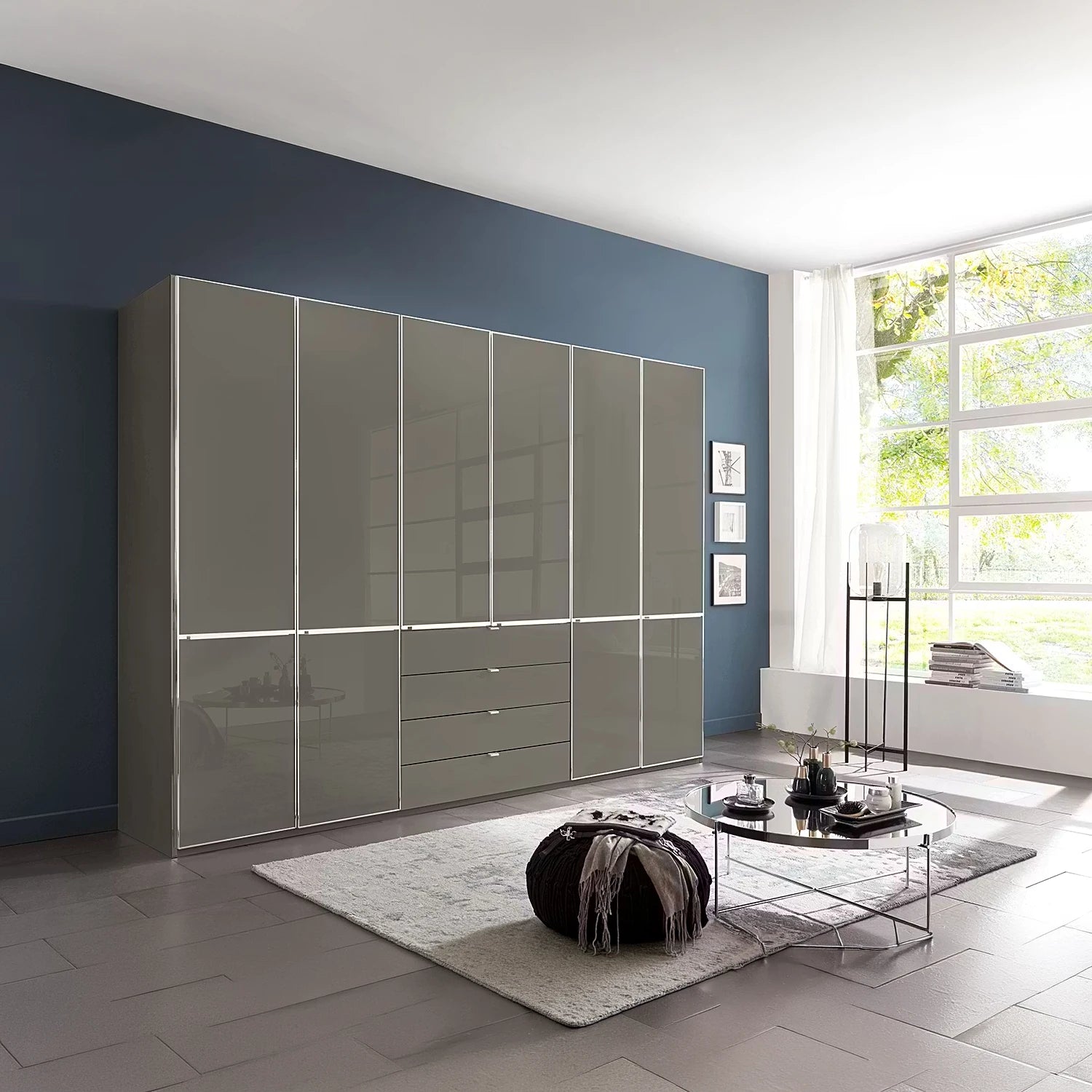 A large modern wardrobe or closet system with glossy Havana Glass doors and drawers, installed against a dark blue wall in a contemporary living room with a window, rug, and coffee table.