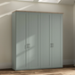 A tall Wiemann Truro four-door wardrobe in a muted sage green color with a natural wood top and silver handles, set against a beige wall with soft window light shadows.