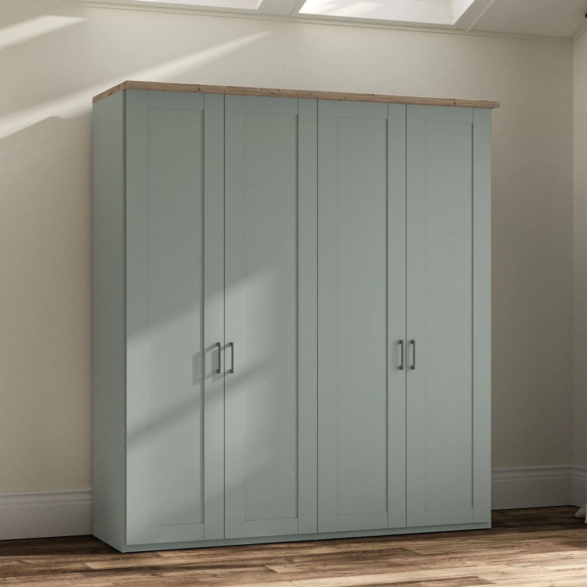A tall Wiemann Truro four-door wardrobe in a muted sage green color with a natural wood top and silver handles, set against a beige wall with soft window light shadows.