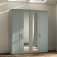 A tall four-door Wiemann truro wardrobe in a muted sage green color with a natural wood top and silver handles, featuring two mirrored doors. It is shown in a real room setting with a wood floor, beige walls, and a curtain visible in the mirror.