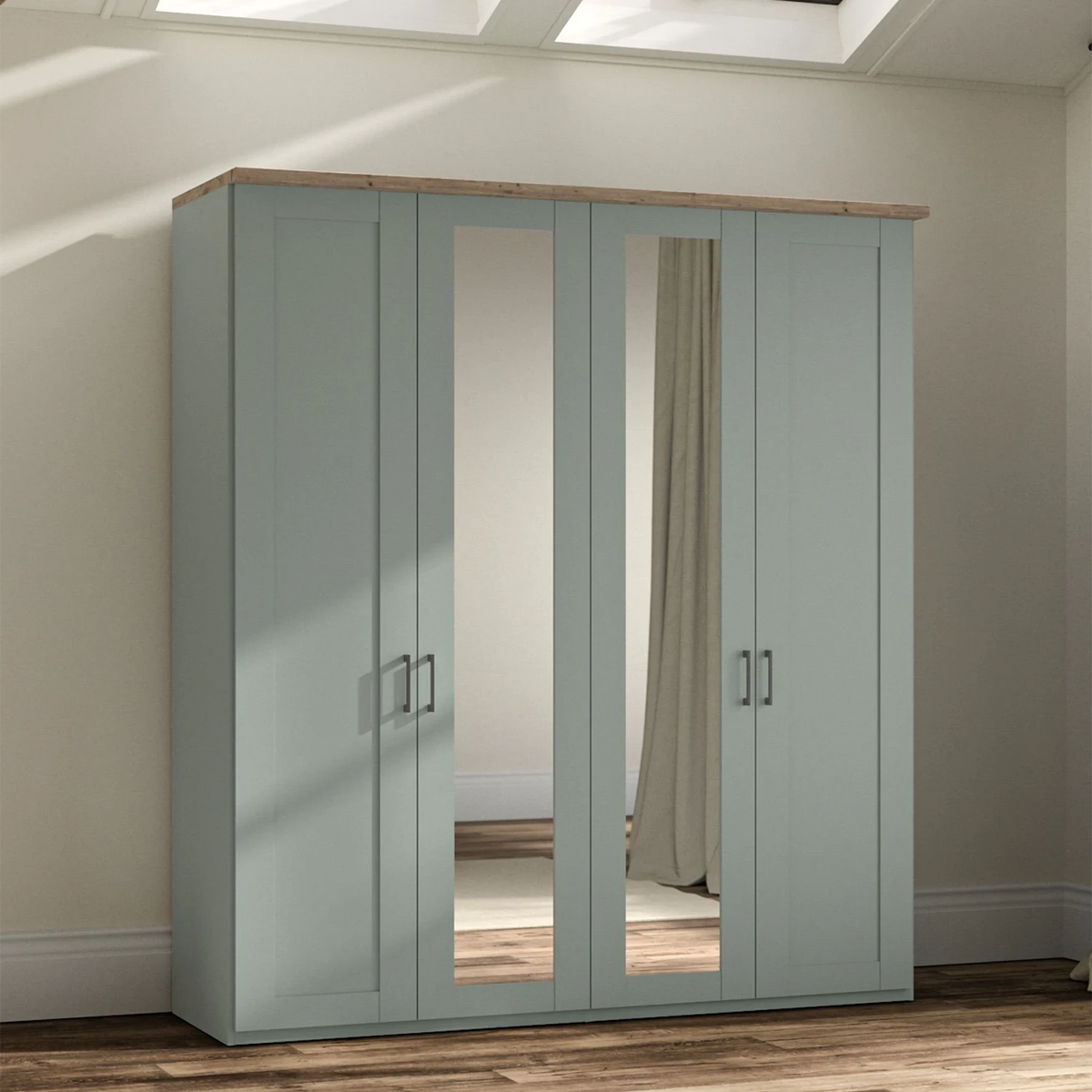 A tall four-door Wiemann truro wardrobe in a muted sage green color with a natural wood top and silver handles, featuring two mirrored doors. It is shown in a real room setting with a wood floor, beige walls, and a curtain visible in the mirror.